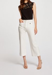 Mujer posando con pantalones blancos cortados y bolsillos frontales, blusa sin mangas de encaje negro y sandalias de tacón alto doradas sobre un fondo liso.