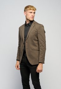Brun blazer i et blød stof, med enkelt knaplukning og to forlommer. Kombineret med en sort skjorte og tætsiddende sorte bukser.