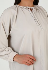 Blusa beige chiaro con scollatura arricciata, chiusura a cordoncino e maniche a sbuffo con polsini abbottonati. Tessuto morbido e design minimale.