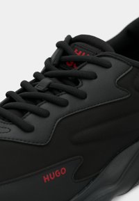 Baskets noires en mesh et caoutchouc, tige texturée, lacets plats, et logo rouge "HUGO" sur le côté.