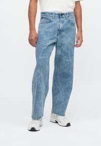 Mężczyzna w niebieskich dżinsach w stylu acid-wash, białych sneakersach i białej koszuli, stojący na tle białego tła.