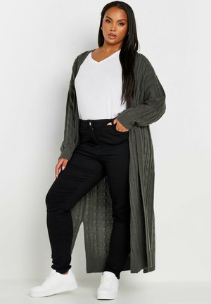 Un largo cárdigan de punto en verde oliva sobre una camiseta blanca con escote en pico, combinado con jeans ajustados negros y zapatillas blancas, mostrando una textura acanalada.