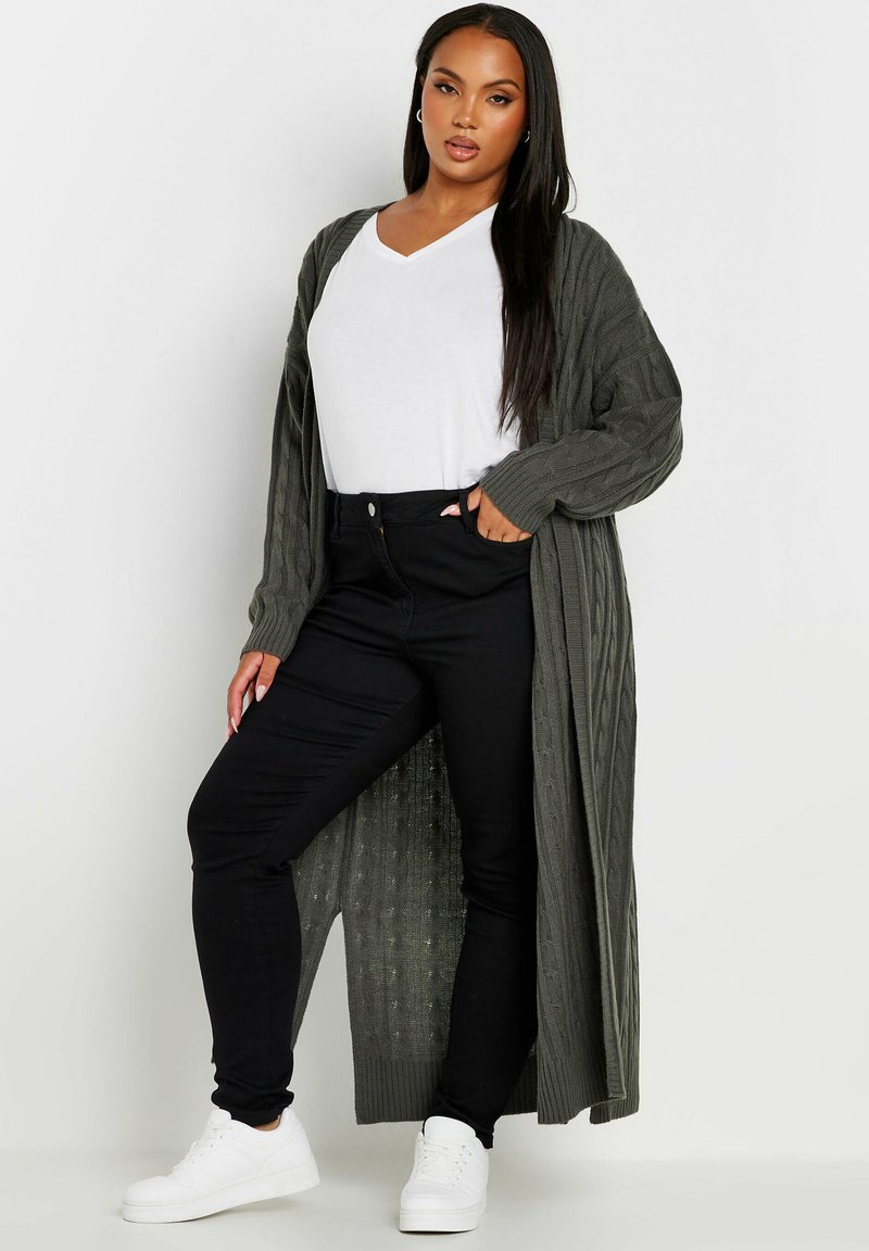 Lång, olivgrön stickad cardigan över en vit v-ringad tröja, kombinerad med svarta skinny jeans och vita sneakers, som visar upp ribbad struktur.