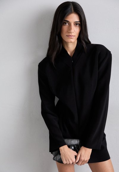 Black long-sleeve top with a high collar, featuring an asymmetric design and a smooth texture. Paired with a metallic clutch bag.