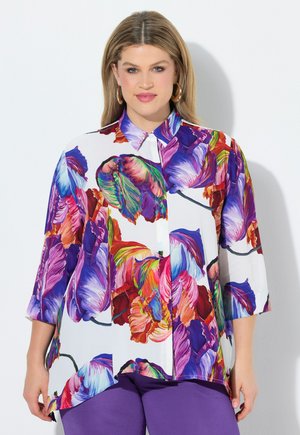 Femme portant une chemise à boutons imprimée de fleurs colorées avec des manches trois-quarts et un pantalon violet, debout sur un fond blanc.