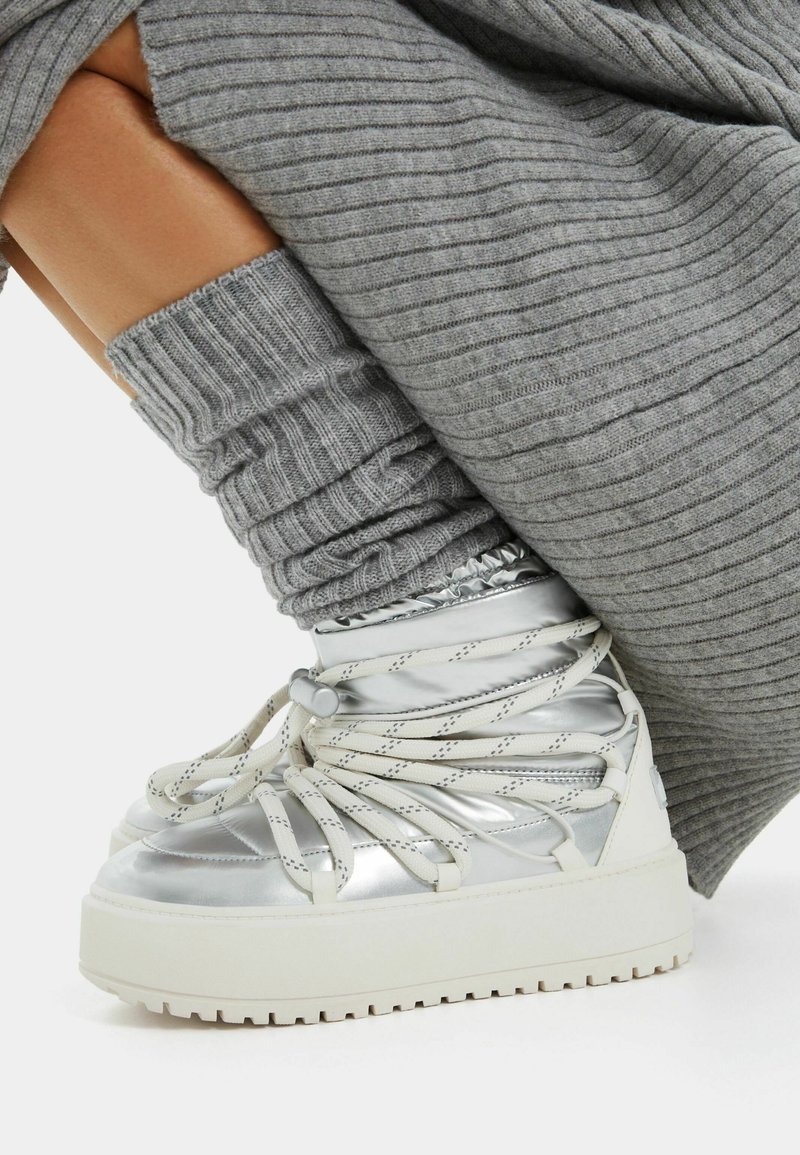 Metallisch silberne High-Top-Sneaker mit weißen Schnürsenkeln, erhöhten weißen Sohlen und strukturierten grauen gestrickten Beinwärmern.