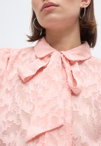 Γυναίκα που φοράει ένα ροζ δαντελένιο πουκάμισο με μεγάλο γιακά με φιόγκο και ασημένια κρίκους σκουλαρίκια, απεικονιζόμενη από τους ώμους έως το πηγούνι.