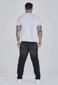 T-shirt grigia a maniche corte, jeans in denim scuro e scarpe da ginnastica bianche; visibili tatuaggi sulle braccia, design semplice senza motivi o logo.