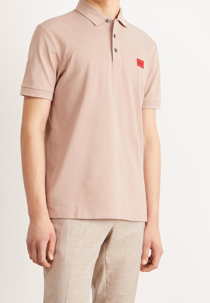 Jasnoróżowa koszulka polo z bawełny, z dwoma guzikami, żebrowanym kołnierzykiem oraz małą czerwoną naszywką na piersi.