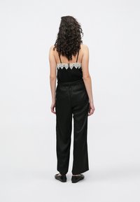 Czarne satynowe spodnie o luźnym kroju, z tyłu zdobione koronkowym wykończeniem na kamizelce oraz regulowanymi cienkimi ramiączkami. Noszone z płaskim obuwiem.