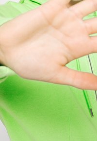 Felpa con cappuccio verde neon con cerniera, realizzata in tessuto morbido e strutturato; tasca frontale visibile; mano che copre parzialmente la vista da destra.