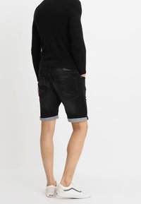 Mustat farkkushortsit, joissa on käännetyt lahkeet, takataskut ja sileä pinta, yhdistettynä valkoisiin lenkkareihin ja mustaan ribbipaitaan.