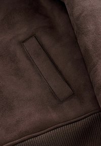 Gros plan sur une poche de veste en daim marron foncé avec des coutures visibles et un ourlet côtelé en bas.