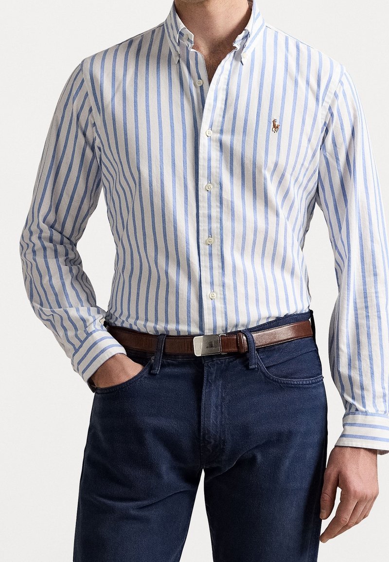 Striped blue and white long-sleeve shirt with a button-down collar, featuring a small logo, paired with dark denim jeans and a brown belt.