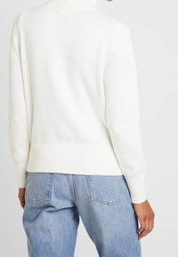 Pull en maille blanche avec un col montant, des poignets et un ourlet côtelés, associé à un jean taille haute bleu clair. La texture semble douce et confortable.