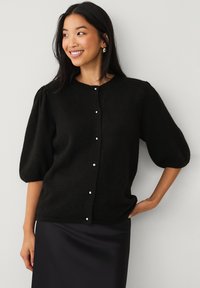 Cardigan lavorato a maglia nero con maniche a palloncino, scollatura rotonda e chiusura frontale con bottoni, caratterizzato da piccoli bottoni bianchi simili a perle. Texture morbida.