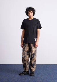 Μαύρο βαμβακερό t-shirt και καφέ cargo παντελόνι tie-dye. Το παντελόνι έχει άνετη εφαρμογή και τσέπες. Το μοντέλο φοράει μαύρα αθλητικά παπούτσια. Απλό φόντο.