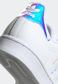Vit lädersneaker med en färgglad iriserande hälflik, texturerad gummisula och "Superstar"-bokstäver i guld.
