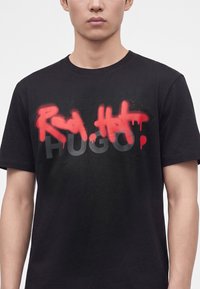 Άνδρας με μαύρο T-shirt με κόκκινο γκράφιτι στυλ σπρέι πάνω από μερικώς ορατό γκρι όνομα επωνυμίας "HUGO" μπροστά.