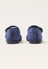 Zapatos azul terciopelo de tipo slip-on con un tacón bajo y un acento decorativo de hebilla dorada en la correa trasera. Textura suave y forma redondeada.