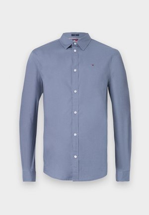 Camicia a maniche lunghe con bottoni in tessuto azzurro chiaro. Presenta un colletto classico, bottoni frontali e un piccolo logo sul petto. Texture liscia.