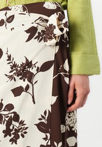 Jupe de style portefeuille avec un fond crème et des motifs floraux marron, nouée à la taille. Haut texturé vert au niveau des manches.