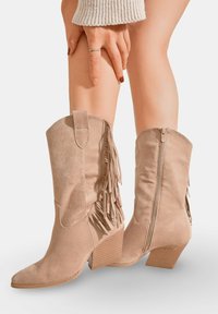 Bottes de cow-boy en daim beige avec des franges, bout pointu et fermeture éclair à l'arrière. Talon block texturé et finition lisse.