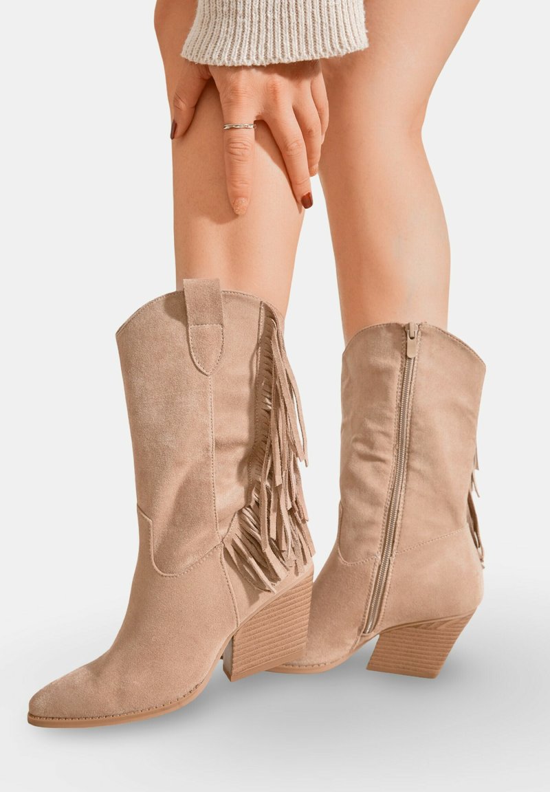 Bottes de cow-boy en daim beige avec des franges, bout pointu et fermeture éclair à l'arrière. Talon block texturé et finition lisse.
