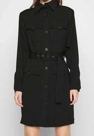 Robe-chemise noire ceinturée avec boutons avant, quatre poches à rabat, manches longues et col large, portée par une personne sur un fond uni.