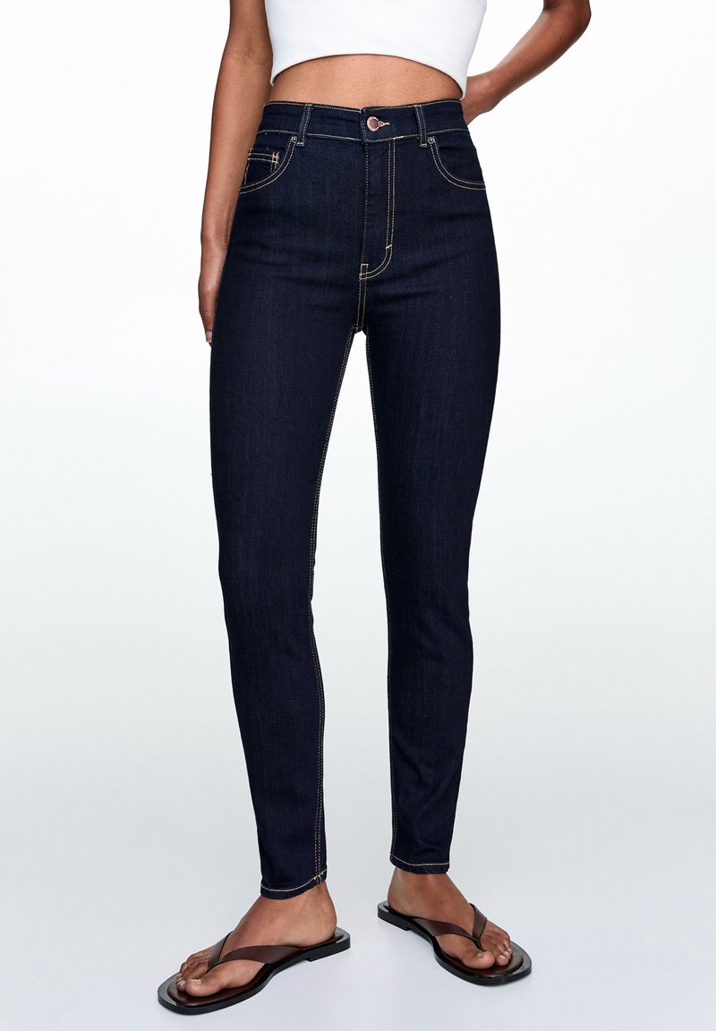 Jeans de tiro alto en azul oscuro con costuras doradas, cinco bolsillos y cierre de botón. Fabricados en denim, con un diseño de ajuste slim.