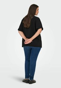 Crna oversized majica s kratkim preokrenutim rukavima, uparena s plavim uskim trapericama. Model je okrenut leđima, ruke su mu povezane iza leđa.