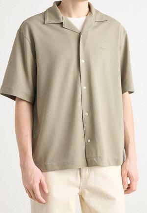 Mand iført en lyse olivengrøn kortærmet skjorte med knapper over en hvid t-shirt med beige bukser, stående mod en ensfarvet baggrund.