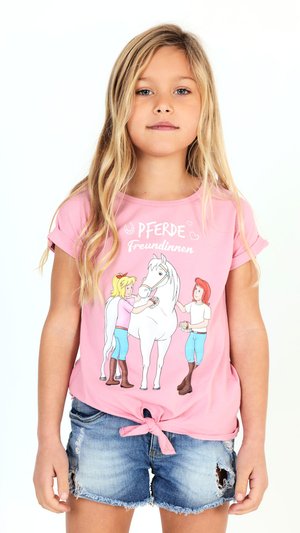 Ragazza bionda che indossa una maglietta rosa con un’illustrazione di un cavallo e due bambine e pantaloncini di jeans strappati, in piedi su uno sfondo bianco.