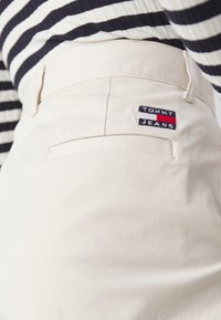 Gros plan sur la poche d'un pantalon beige avec un patch logo Tommy Jeans, porté avec un haut rayé noir et blanc.