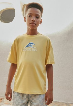 Junge Person steht drinnen und trägt ein gelbes "Catch Some Waves" T-Shirt und karierte Shorts vor einer weißen strukturierten Wand.