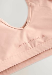 Mjukt rosa sport-bh med "NEXT STUDIO"-logotyp prägla på framsidan, med ett brett elastiskt band och sömlös design.