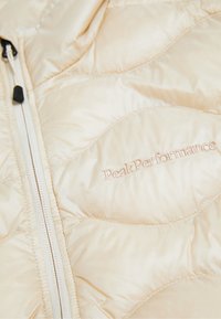 Beige Pufferjacke mit gestepptem Design, ausgestattet mit einem Frontzipper und dem aufgestickten "Peak Performance"-Logo in heller Fadens. Glatte Stoffstruktur.