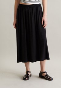 Sort, ankel-lang nederdel med en indsamlet talje, lavet af blødt stof. Bæres sammen med sorte sandaletter med stropper, der har et enkelt design.