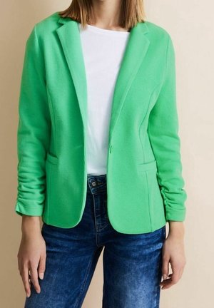 Personne portant un blazer vert vif sur une chemise blanche et un jean bleu, debout devant un fond uni beige clair.