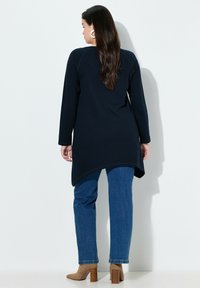 Marineblauwe tuniek met lange mouwen en een afgeronde zoom, gecombineerd met blauwe recht gesneden spijkerbroek en beige enkellaarsjes met blokhak. Gladde stoftextuur.