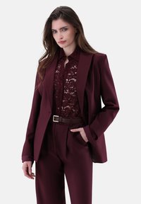 Blazer color borgogna e pantaloni su misura, abbinati a una blusa in pizzo borgogna. Presenta una cintura con fibbia argentata e un design in tessuto texture.