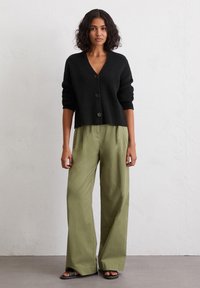 Schwarze Strickjacke mit V-Ausschnitt und drei Knöpfen, kombiniert mit lockeren, hellolivgrünen Hosen. Glatte Textur und lässige Passform.