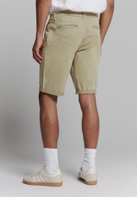 Beige katoenen shorts met omgeslagen zomen, twee achterzakken en een zijzak. Geddragen met witte sokken en beige sneakers op een neutrale achtergrond.