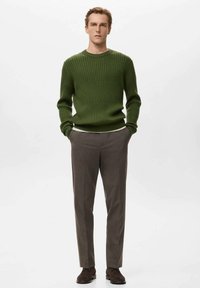 Zelený rebrovaný sveter s okrúhlym výstrihom, doplnený hnedými strihanými nohavicami. Model nosí tmavé topánky a stojí na jednoduchom pozadí.