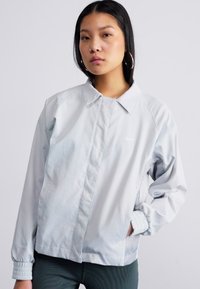 Lys grå Nike-jakke med krage, lange ermer, elastiske mansjetter og en glatt tekstur. Minimalistisk design uten synlige mønstre.