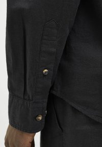 Camicia nera di lino con maniche lunghe, caratterizzata da un polsino con bottoni e dettagli delle cuciture verticali. Il tessuto ha una finitura texturizzata e una leggera lucentezza.