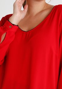 Kobieta w czerwonej bluzce z długim rękawem i dekoltem w kształcie litery V, z czarnymi wykończeniami, dotyka szyi prawą dłonią w pobliżu twarzy.