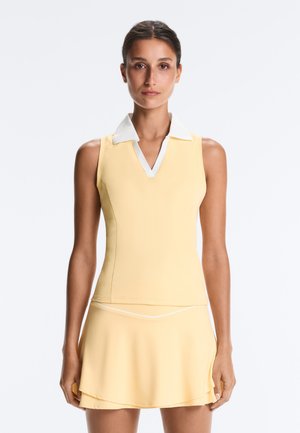 Femme portant un haut sans manches jaune pâle à col et une jupe assortie, debout devant un fond blanc uni.