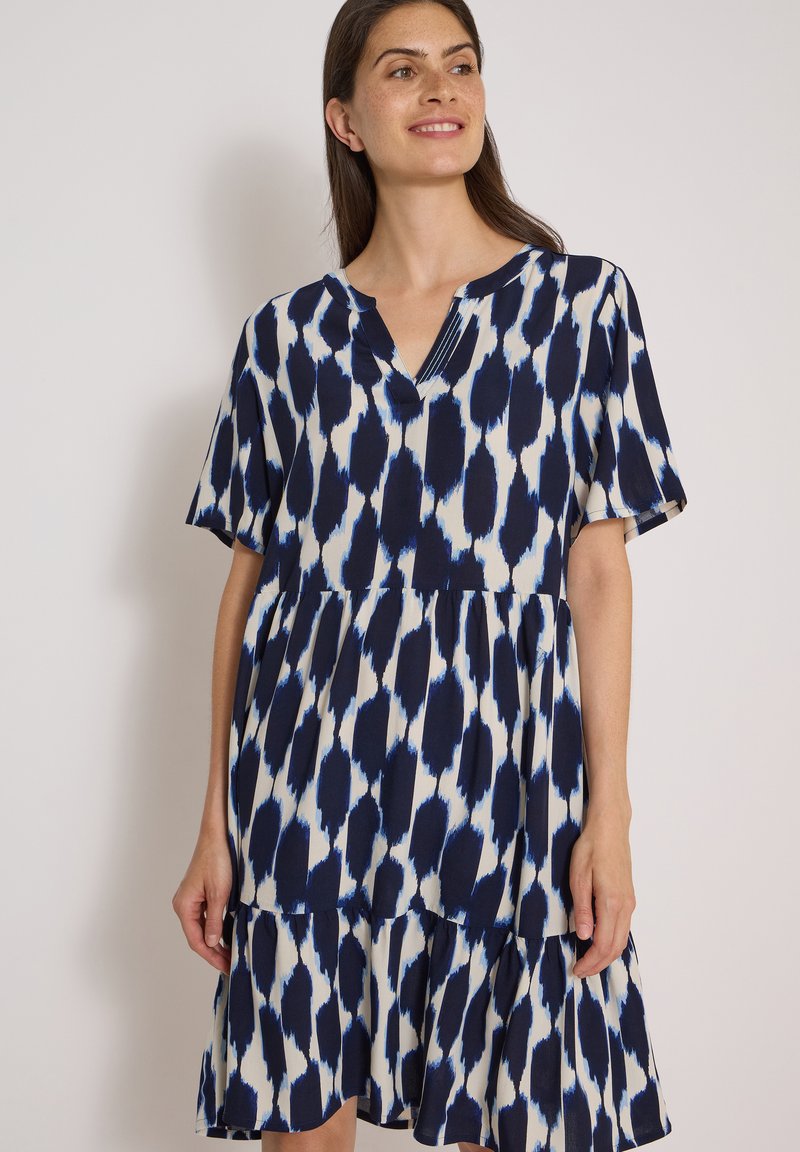 Femme portant une robe ample à manches courtes avec un motif abstrait bleu et blanc, debout devant un fond clair uni.