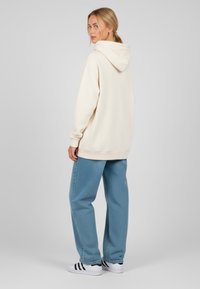 Felpa oversize color crema con cappuccio regolabile, abbinata a pantaloni di jeans blu. Sneakers bianche con strisce nere. Tessuto morbido, vestibilità comoda.